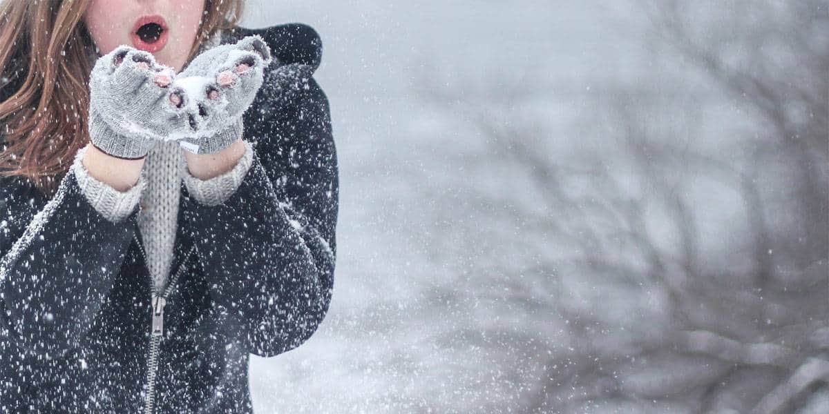 Девојка во зимска јакна и сиви ракавици дува снег од дланките